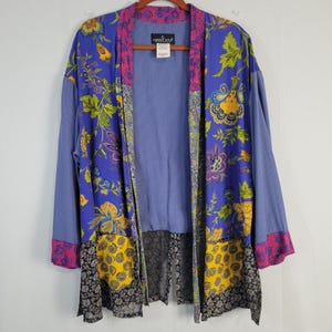 May include: A long-sleeved open-front cardigan featuring a vibrant blue base with a multi-colored floral and paisley print. The garment includes panels of yellow, black, and pink patterns, with a textured light blue interior and a Carole Little label.