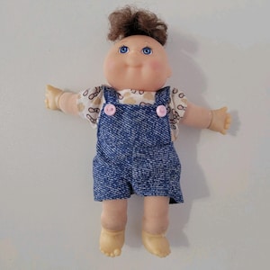 May include: A doll with a round head, brown hair, and blue eyes. It wears a short-sleeved shirt with a tan and brown pattern under blue denim-style overalls. The overalls feature two small pink buttons at the straps. The doll has light skin.