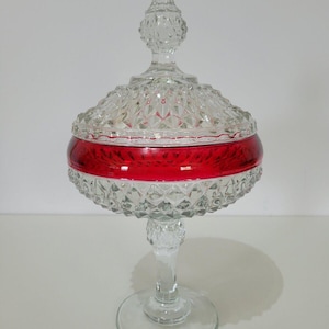 May include: A clear glass candy dish with a red band around the middle. The dish has a diamond pattern and a lid with a pointed finial. The base is a clear glass pedestal.
