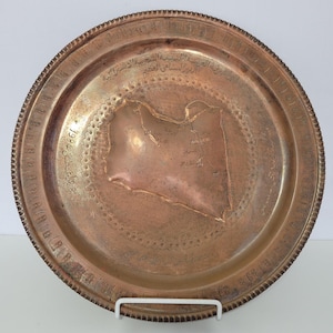 May include: A round copper plate featuring an embossed map of Libya in the center. The plate includes engraved Arabic text along the rim and across the map surface, with the date 1984 visible on the left side. The edge has a decorative scalloped border.