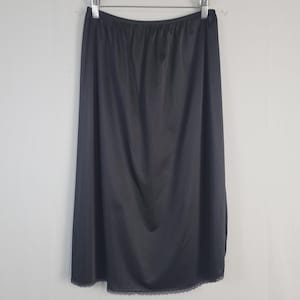 May include: A black slip skirt hanging on a hanger. The skirt is made of a smooth, shiny fabric and has a lace trim at the hem. The skirt is a solid black color.