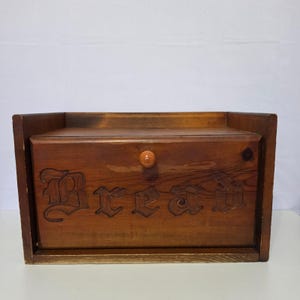 May include: A rectangular wooden bread box with a medium brown finish and a visible wood grain. The front panel features the word Bread carved in a decorative font and includes a small, round, light-colored wooden knob for opening the door.
