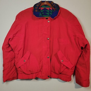 May include: A bright red jacket with a patterned collar featuring blue, green, and brown geometric shapes. The jacket has two front pockets, a button-up closure, and a ribbed hem. The jacket is a vintage style.