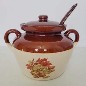 May include: A two-toned ceramic jar with a lid and spoon. The top and lid are brown, while the bottom is cream-colored with a fruit and nut design. The jar has two handles and a spoon resting on the side.