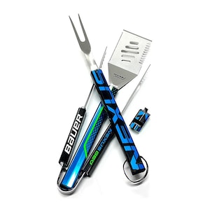 May include: A set of stainless steel grilling tools with blue and black handles. The handles feature the logos "Bauer" and "Nexus Geo". The set includes a spatula, fork, and tongs. The tools are designed to look like hockey sticks.