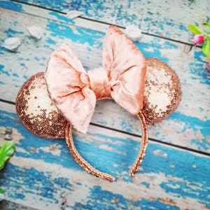 May include: Rose gold sequined mouse ears headband with a large velvet bow. The headband is a popular accessory for theme park visits.