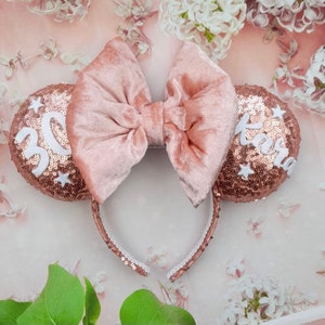 May include: Rose gold sequin Minnie Mouse ears headband with a large velvet bow. The left ear has the number 30 and white stars, the right ear has the name "Tara" and white stars.