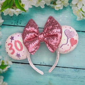 May include: White Minnie Mouse ears with a pink sequin bow, a white ear with the number 30 and pink stars, and a white ear with pink hearts and balloons.