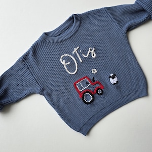 May include: A blue long-sleeve knit jumper featuring the name Otis embroidered in white yarn. Below the name is a red tractor with black wheels and a small white and black sheep figure, all rendered in a textured, raised yarn embroidery style.