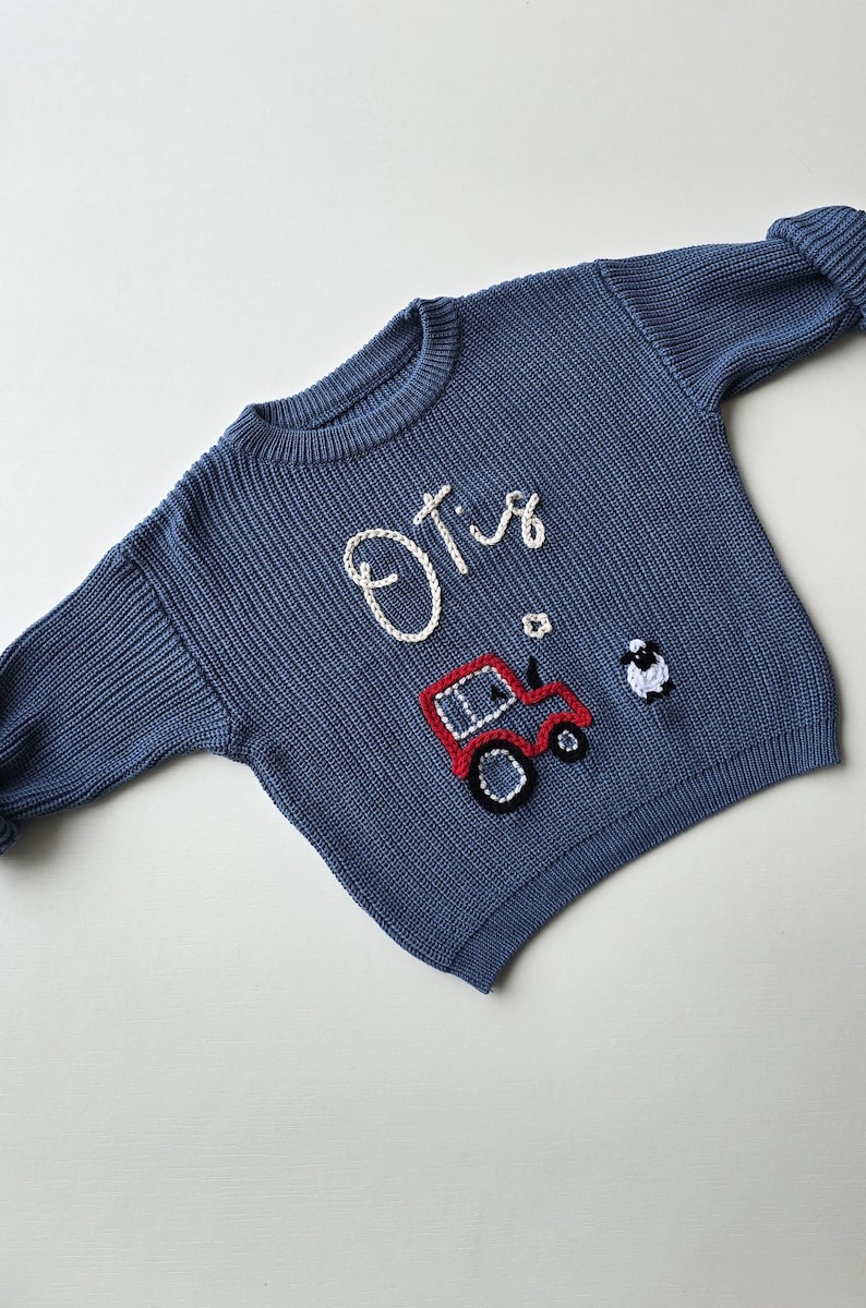 May include: A blue long-sleeve knit jumper featuring the name Otis embroidered in white yarn. Below the name is a red tractor with black wheels and a small white and black sheep figure, all rendered in a textured, raised yarn embroidery style.