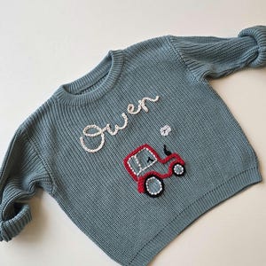 May include: A dusty blue long-sleeve knit jumper featuring the name Owen embroidered in white cursive script above a red tractor graphic with black and white detailing. The jumper has a ribbed crew neckline, cuffs, and hem.