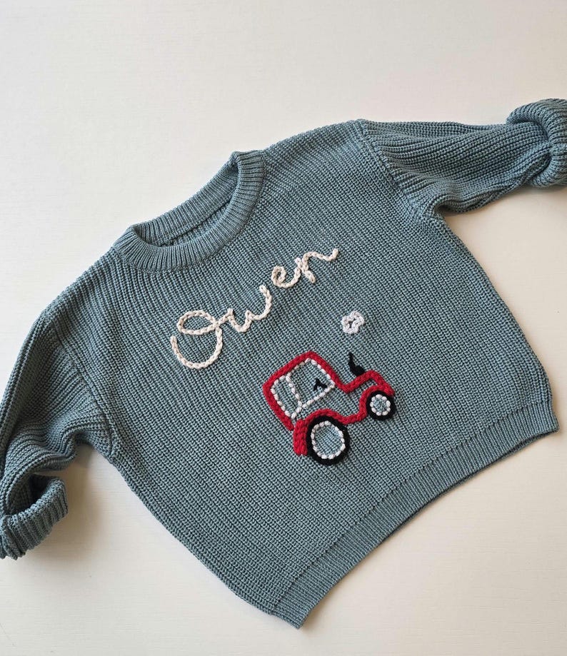 May include: A dusty blue long-sleeve knit jumper featuring the name Owen embroidered in white cursive script above a red tractor graphic with black and white detailing. The jumper has a ribbed crew neckline, cuffs, and hem.