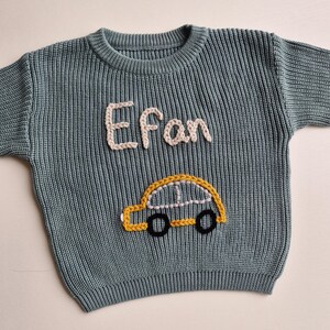 May include: A sage green long-sleeve knit jumper featuring a crew neckline and ribbed texture. The front displays the name Efan embroidered in cream-coloured yarn above a yellow car graphic with black wheels and white window detailing.