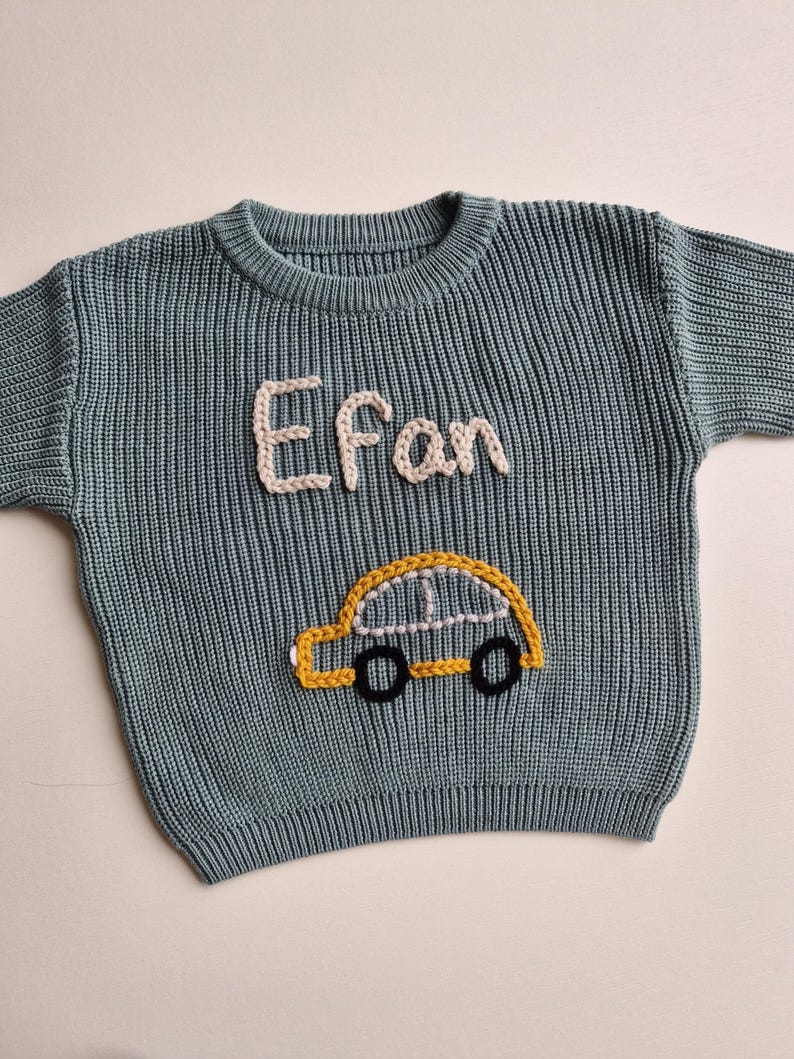 May include: A sage green long-sleeve knit jumper featuring a crew neckline and ribbed texture. The front displays the name Efan embroidered in cream-coloured yarn above a yellow car graphic with black wheels and white window detailing.