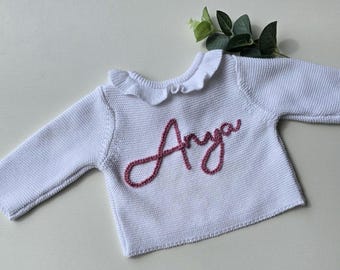 May include: White baby sweater with long sleeves and a frilled collar. The name "Anya" is embroidered in pink cursive across the chest. A sprig of greenery is in the background. This knitted sweater is perfect for a baby.