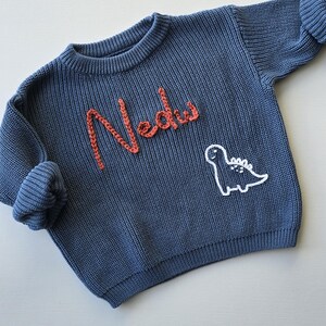 May include: A blue long-sleeve knit jumper featuring the name Nedu embroidered in orange yarn on the chest. Below the name, a white outline of a dinosaur is stitched into the fabric. The jumper has a ribbed crew neckline and ribbed cuffs.