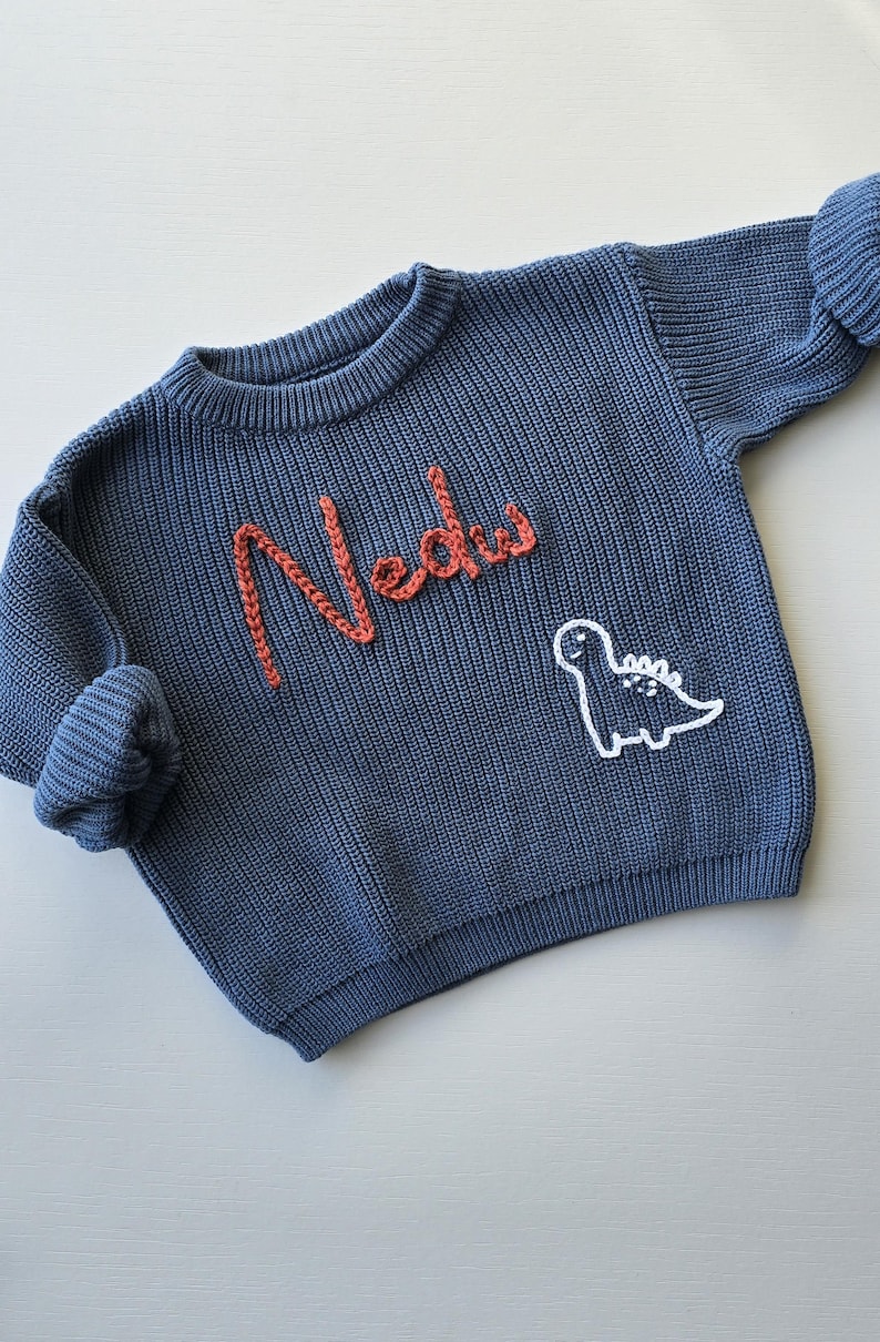 May include: A blue long-sleeve knit jumper featuring the name Nedu embroidered in orange yarn on the chest. Below the name, a white outline of a dinosaur is stitched into the fabric. The jumper has a ribbed crew neckline and ribbed cuffs.
