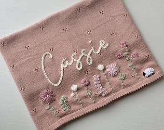 May include: A dusty pink knitted blanket featuring a pointelle pattern and the name Cassie embroidered in white yarn. The blanket is decorated with embroidered flowers in shades of pink, white, and green, and a small white sheep with a black face.