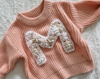 May include: A pale pink knitted jumper with a large white, beige and cream crocheted letter "M" on the front.