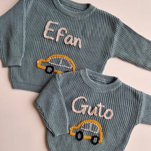 May include: Two blue-grey long-sleeve knit jumpers featuring embroidered details. One jumper displays the name Efan in light pink yarn above a yellow car with black wheels. The other jumper displays the name Guto in light pink yarn above a yellow car.