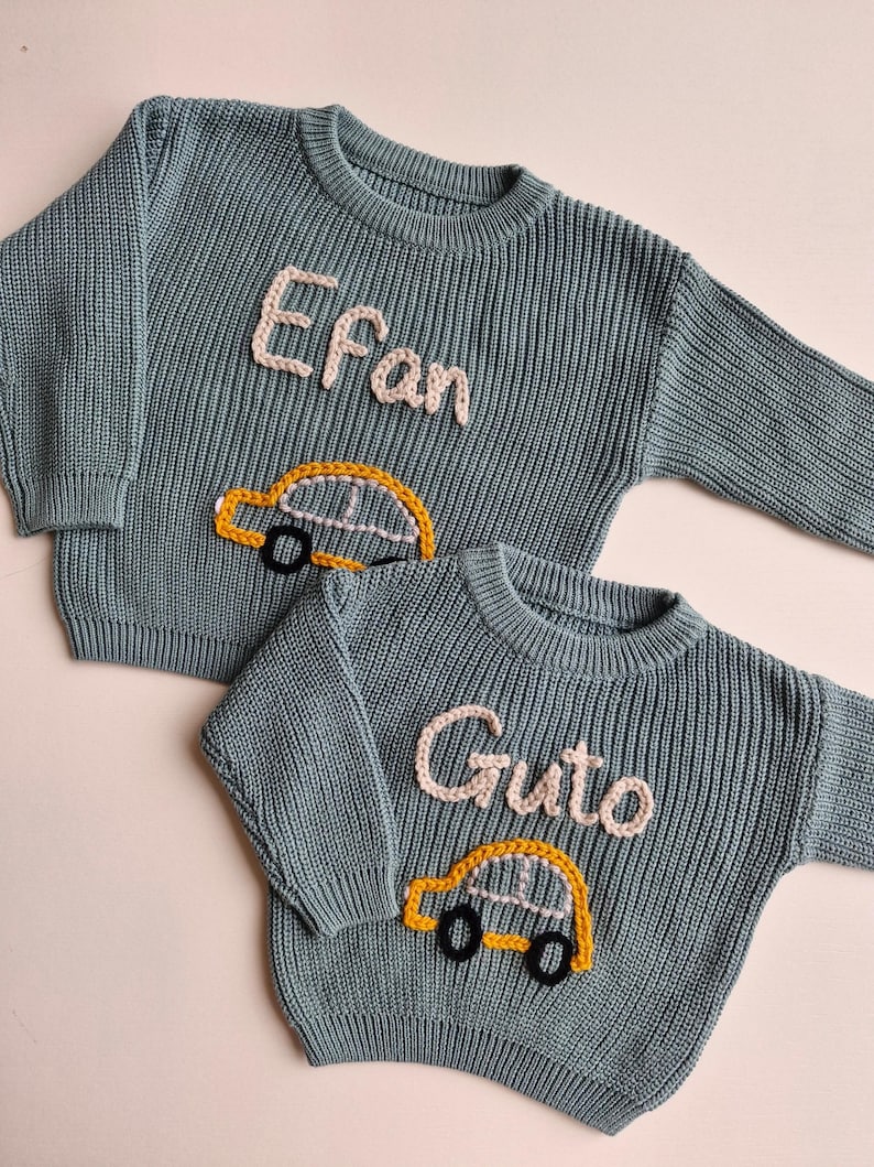 May include: Two blue-grey long-sleeve knit jumpers featuring embroidered details. One jumper displays the name Efan in light pink yarn above a yellow car with black wheels. The other jumper displays the name Guto in light pink yarn above a yellow car.