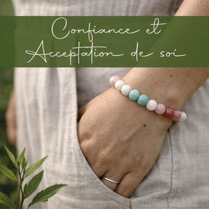 Può includere: Un braccialetto di perline con pietre rotonde e lisce nei toni del bianco, rosa pallido, turchese e rosso intenso. Il braccialetto è indossato al polso e un banner verde in alto mostra il testo corsivo bianco Confiance et Acceptation de soi.