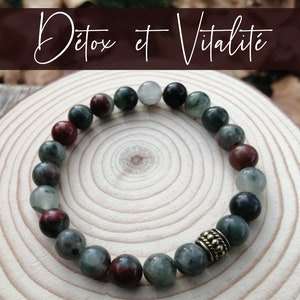 May include: A green, red and black beaded bracelet with a gold charm. The bracelet is on a wooden surface. The text "Detox et Vitalité" is in the background.