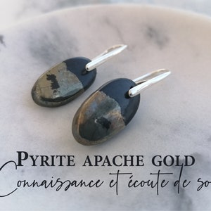 Può includere: Un paio di orecchini d'argento con pietre ovali di pirite e oro apache. Le pietre sono nere con macchie dorate. Il testo "PYRITE APACHE GOLD Connaissance et écoute de soi" è visibile nell'immagine.