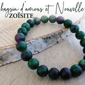 Puede incluir: Un brazalete de piedra zoisita verde y rosa con el texto "Chagrin d'amour et Nouvelle vie" y "ZOÏSITE" sobre una superficie de madera.