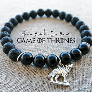 Könnte beinhalten: Armband aus schwarzen Lavasteinen und Onyxperlen mit einem silbernen Wolfsanhänger. Das Armband trägt den Text "House Stark. Jon Snow. GAME OF THRONES".