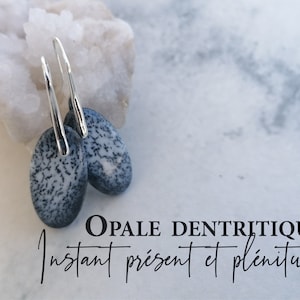Peut inclure: Une paire de boucles d'oreilles pendantes en argent avec des pierres d'opale dendritique de forme ovale. Les pierres sont un mélange de blanc et de gris avec des motifs dendritiques noirs. Les boucles d'oreilles sont sur un fond blanc avec un cristal de roche derrière elles. Le texte "OPALE DENTRITIQUE Instant présent et plénitude" est sous les boucles d'oreilles.