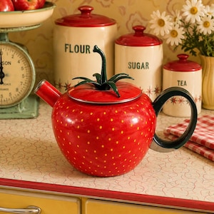 Puede incluir: Una tetera de cerámica de color rojo brillante con forma de fresa, pequeños puntos amarillos, un asa en la tapa con forma de tallo verde y un mango curvo de color verde oscuro. La tetera tiene un pico corto y un cuerpo redondeado.