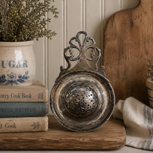 Puede incluir: Un colador de té de metal plateado con un cuenco circular, un centro perforado y un mango ornamental con un diseño de recortes en espiral. El colador presenta un acabado desgastado y una forma clásica y funcional.