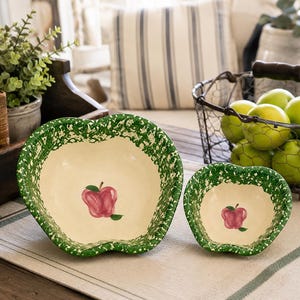May include: Two apple-shaped ceramic bowls with a cream-colored interior and a green speckled rim. Each bowl features a small, hand-painted red apple illustration with a green leaf at the center of the basin. The bowls have a distinct, curved silhouette.