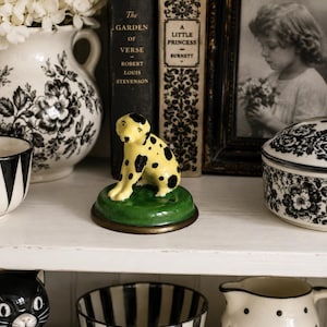 May include: A small ceramic figurine of a yellow dog with black spots, sitting on a circular green base. The dog has a smooth, glossy finish and is positioned in the center of the frame, surrounded by various decorative household items.