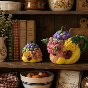 Puede incluir: Una tetera de cerámica y un azucarero a juego con tapa, ambos con un diseño en relieve tridimensional y colorido de varias frutas, incluyendo uvas moradas, piñas amarillas y frambuesas rosas, con hojas verdes y una base amarilla.