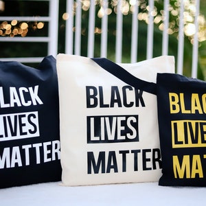 May include: Three tote bags with the phrase "Black Lives Matter" printed on them. The bags are black, white, and yellow.