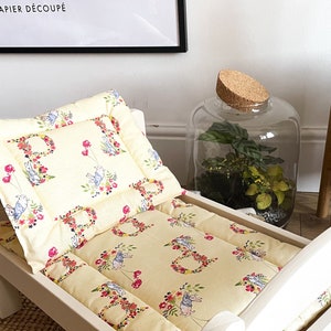 Op de afbeelding: Een wit houten poppenbed met een gele, met bloemen bedrukte matras en kussen. Het kussen heeft een bloemenkransontwerp met een konijn in het midden. Het bed staat op een lichtbruin tapijt.