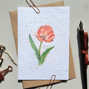 May include: A watercolour painting of a single orange tulip on a white card with a speckled texture. The card is surrounded by a brown envelope and a brown pen with a copper-coloured ribbon.