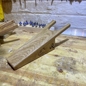 May include: A wooden wedge with a handle, possibly for carpentry. The light-coloured wood has a prominent grain. The initials "W+W" are visible on the side. The wedge is on a wooden workbench with other tools and wood shavings.