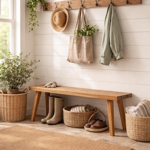 May include: A wooden wall-mounted coat rack with black hooks holding a straw hat, a beige canvas tote bag, and a sage green jacket. Below, a wooden bench sits above three woven baskets, a pair of beige boots, and brown leather shoes.