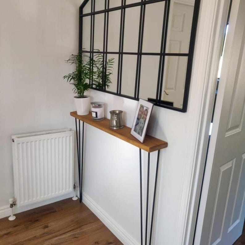 K&ouml;nnte beinhalten: Ein Konsolentisch aus Holz mit schwarzen Haarnadelbeinen. Der Tisch ist mit einer Topfpflanze, Kerzen und einem gerahmten Foto dekoriert. Der Tisch steht an einer wei&szlig;en Wand mit einem schwarzen Spiegel dar&uuml;ber.