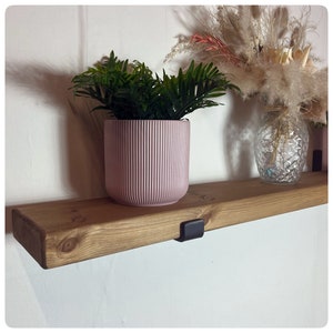 May include: A wooden shelf with a light brown finish, supported by black brackets, holds a pink, ribbed planter with a green plant. A clear glass vase with dried flowers sits to the right. The shelf is mounted on a white wall.