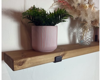 May include: A wooden shelf with a light brown finish, supported by black brackets, holds a pink, ribbed planter with a green plant. A clear glass vase with dried flowers sits to the right. The shelf is mounted on a white wall.