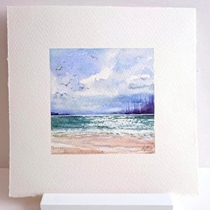 Puede incluir: Pintura acuarela de un paisaje marítimo con una playa de arena, agua azul y un cielo nublado. Una línea de árboles es visible en la distancia.