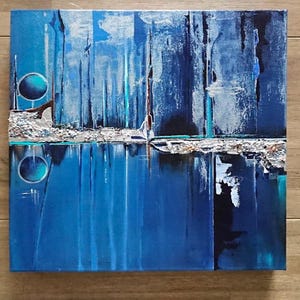 Op de afbeelding: Abstract schilderij op canvas met blauwe, zilveren en witte verf. Het schilderij heeft een horizontale lijn in het midden, met een reflectie van de bovenste helft in de onderste helft. De bovenste helft heeft een getextureerd, abstract ontwerp met een cirkel in de linkerbovenhoek. De onderste helft heeft een blauwe, waterige reflectie.