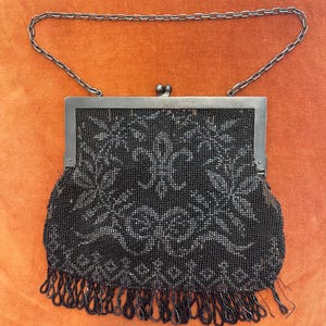 May include: A black beaded handbag featuring a floral pattern and a bottom fringe of beaded loops. The bag has a dark metal frame with a kiss-lock closure and a thin matching metal chain strap for carrying over the shoulder or by hand.
