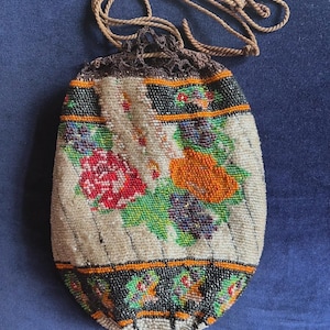 May include: A drawstring pouch featuring intricate beadwork in a floral pattern with red, orange, and purple flowers on a cream background. The bag has black beaded borders with orange accents, a brown crocheted top edge, and a brown twisted rope cord.