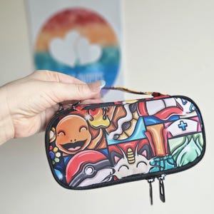 May include: A rectangular zipped carrying case featuring a colourful graphic print of various cartoon characters, including a smiling orange creature, a red and white ball, and a cat-like figure, with black trim and two black zip pulls.