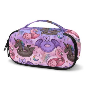 May include: A purple and pink cosmetic bag with a black zipper and handle. The bag features a repeating pattern of inflatable unicorns, flamingos, and bats. The design includes moons, stars, and spiderwebs.